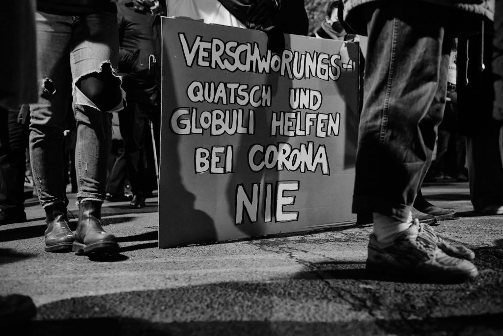 Schild mit der Aufschrift „Verschwörungsquatsch und Globuli helfen bei Corona nie!“ steht inmitten zahlreicher Beine von Demonstrierenden auf dem Boden.